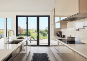 Modern kitchen with white marble counters and an island, stainless appliances, and large black-framed glass doors opening to a sunny countryside view.