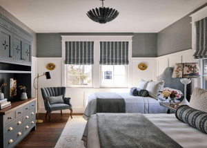 Blue-gray bedroom with two beds, patterned window valances, white wainscoting, and built-in blue cabinets on the left under a black pendant chandelier.