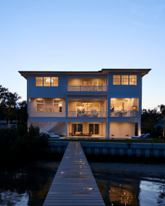 Blue, three-story waterfront home with warm interior lighting visible through large windows and a long wooden dock extending toward the viewer at dusk.