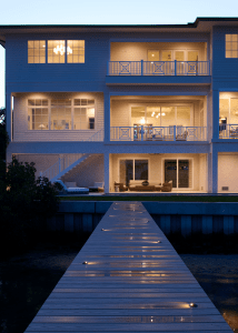 Evening view of a modern white apartment building with illuminated interiors and a long wooden boardwalk leading to the water.