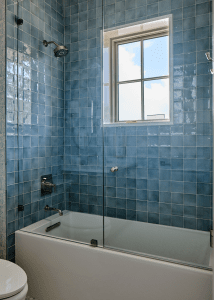 Blue-tiled bathroom with a glass shower enclosure, chrome fixtures, and a window letting in daylight. A toilet is visible in the foreground.