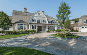 Large cream-colored house with a steep gabled roof, green shutters, and a circular stone driveway bordered by flowering beds and trees on a sunny day.