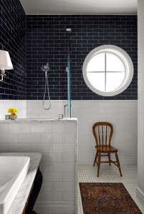 Bathroom with black subway tile walls, a glass shower enclosure, round window, marble countertop, and a wooden chair nearby.