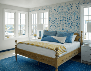 Bedroom interior with a wooden bed, blue patterned wallpaper, and blue accent pillows by large windows; dotted blue rug on the floor.
