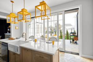 Bright kitchen with a white island, wooden cabinetry, and three large yellow pendant lantern lights overhead, opening to a sunny backyard through glass doors.