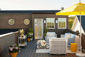 Outdoor wooden deck with wicker furniture, a bright yellow umbrella, and potted plants on a sunny terrace.