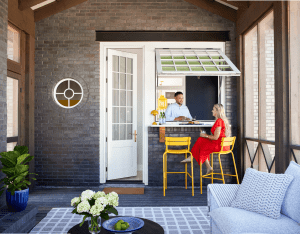 Two people converse at an open kitchen window on a brick porch, bright yellow stools nearby.